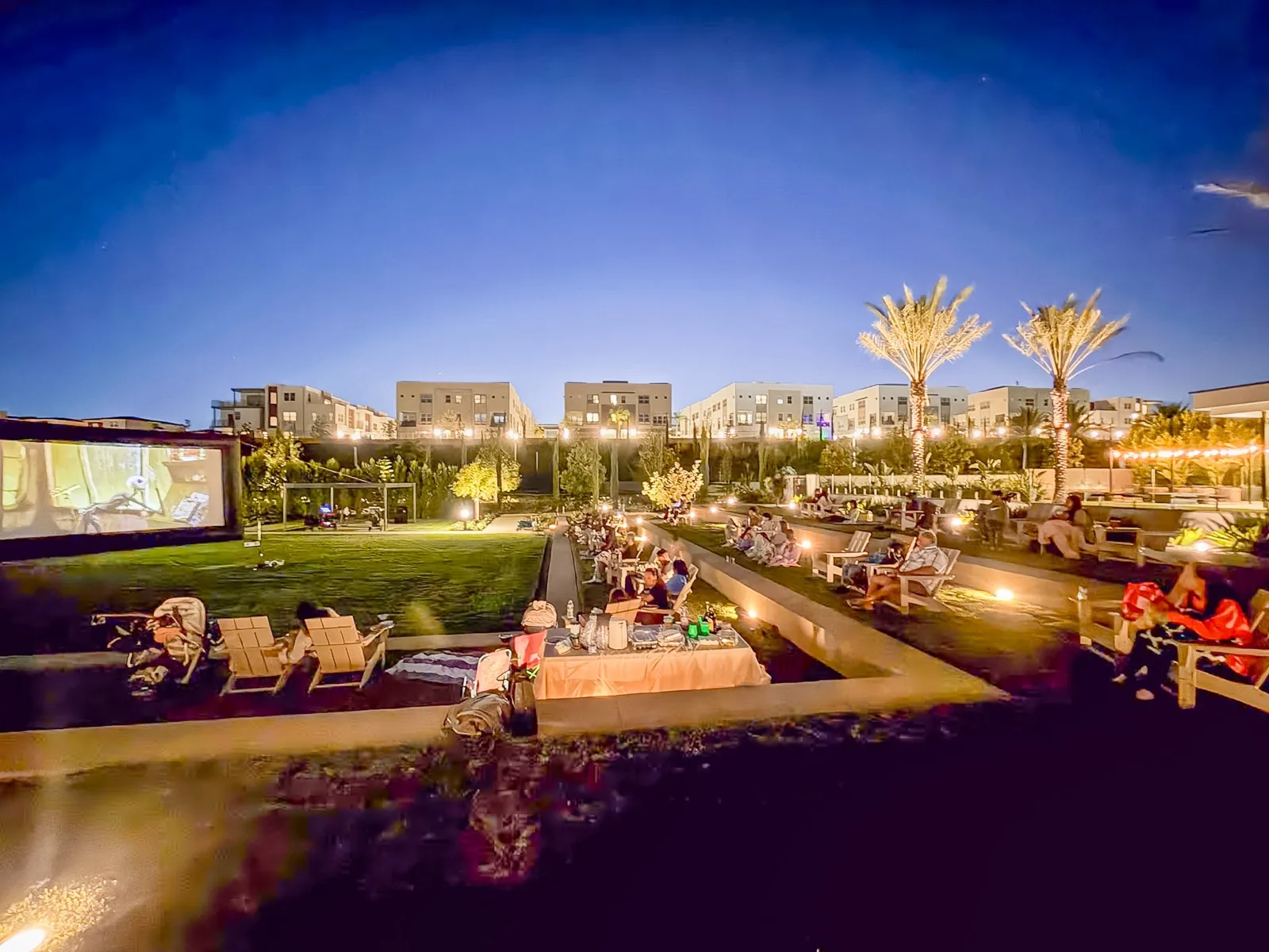 Movie night on the clubhouse lawn at blue hour, string lights on, neighbors in lawn chairs facing the screen.