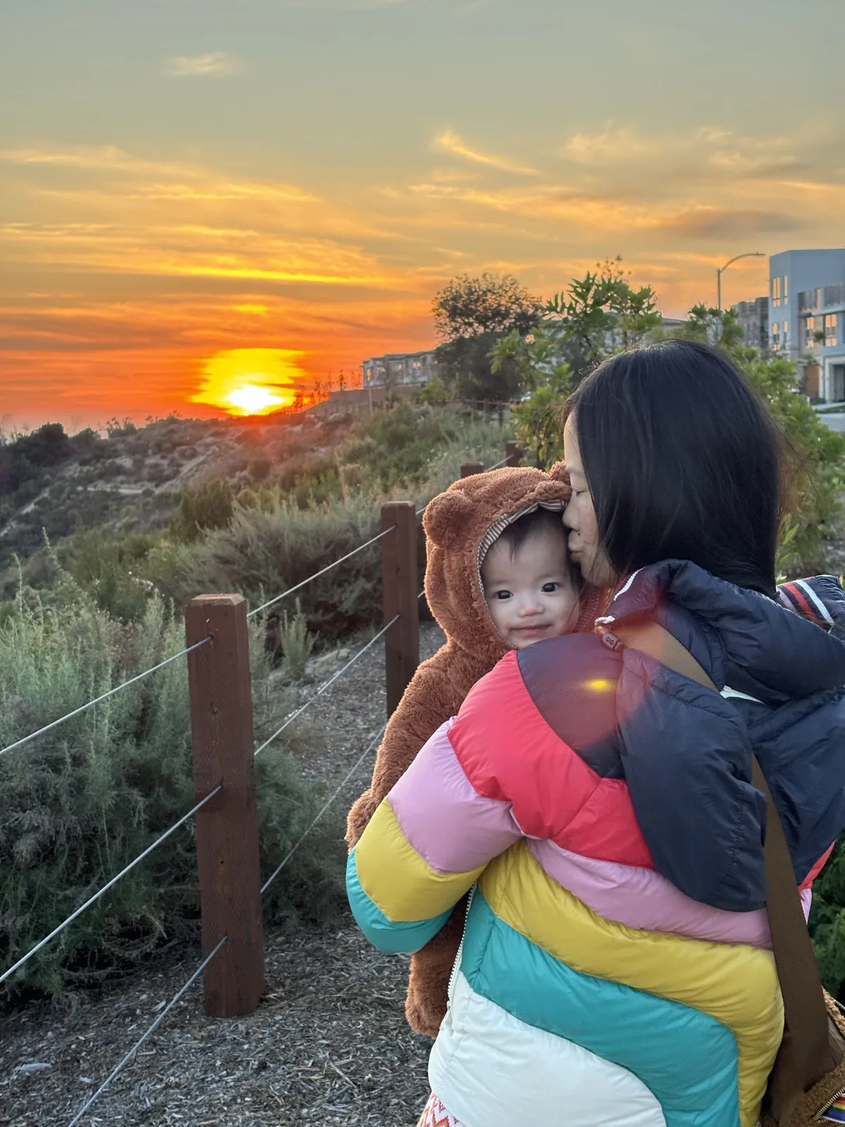 A woman holding a baby on the ridge trail at sunset. The sun sits on the basin behind them, and the Metro Heights homes are on the hill beyond the trail fence.